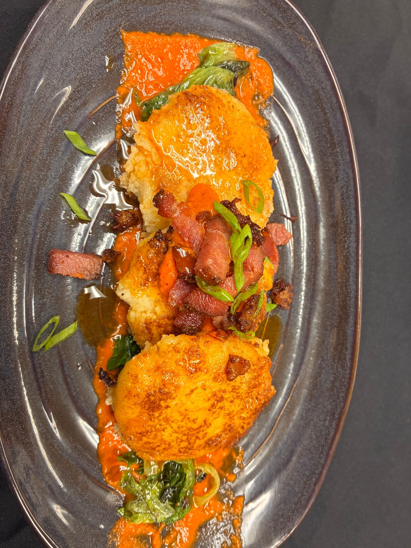 Three golden rounds with diced browned garnish, sliced green onions, greens and orange sauce on a dark oval plate
