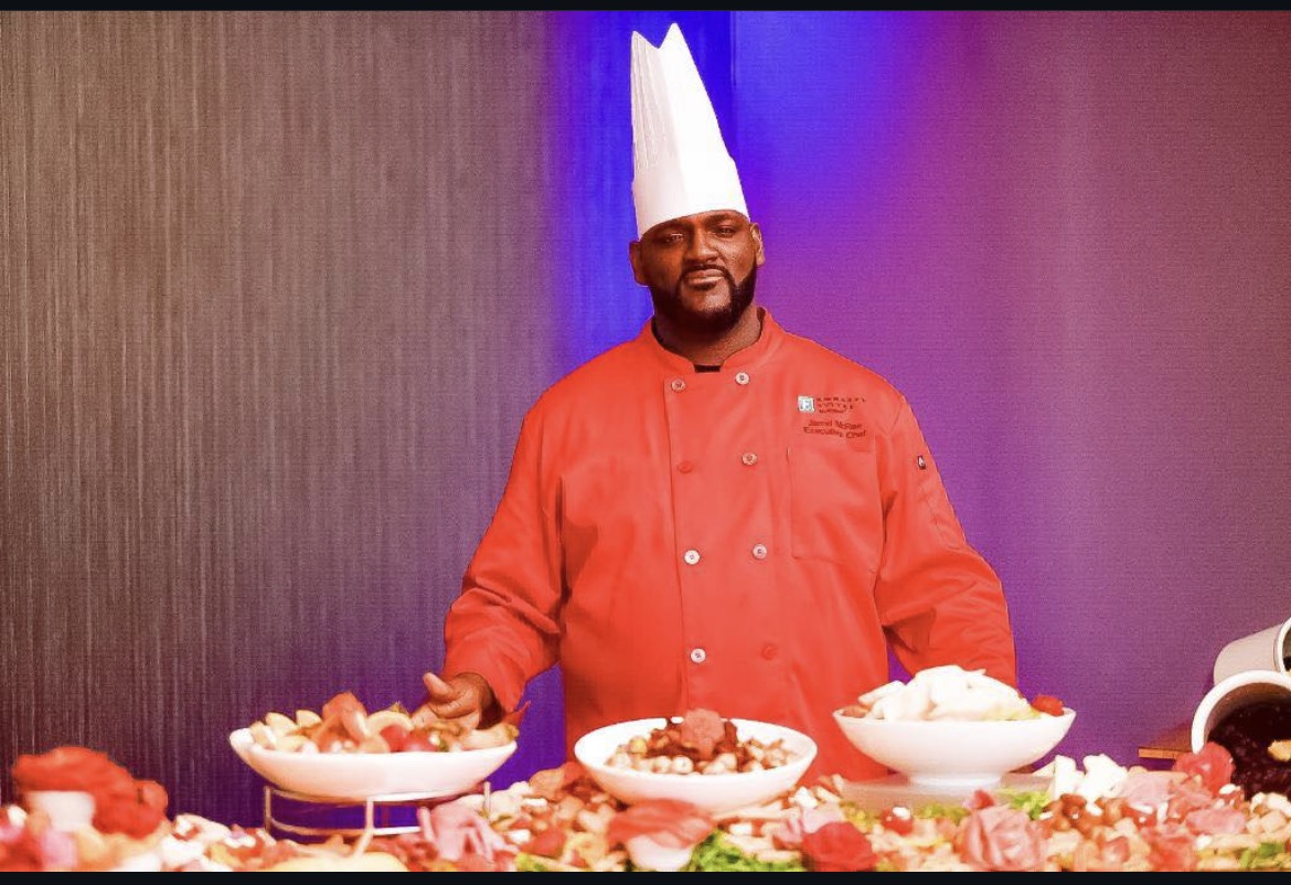 Chef Jarrel McRae in a red chef coat and white toque behind a food display